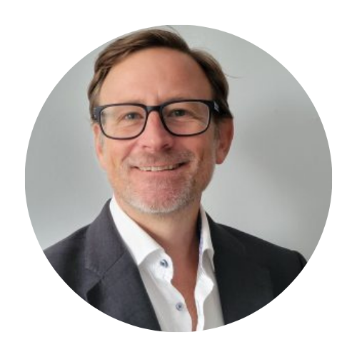 Professional headshot of a smiling man wearing glasses, a dark blazer, and a white shirt against a neutral background for a corporate profile