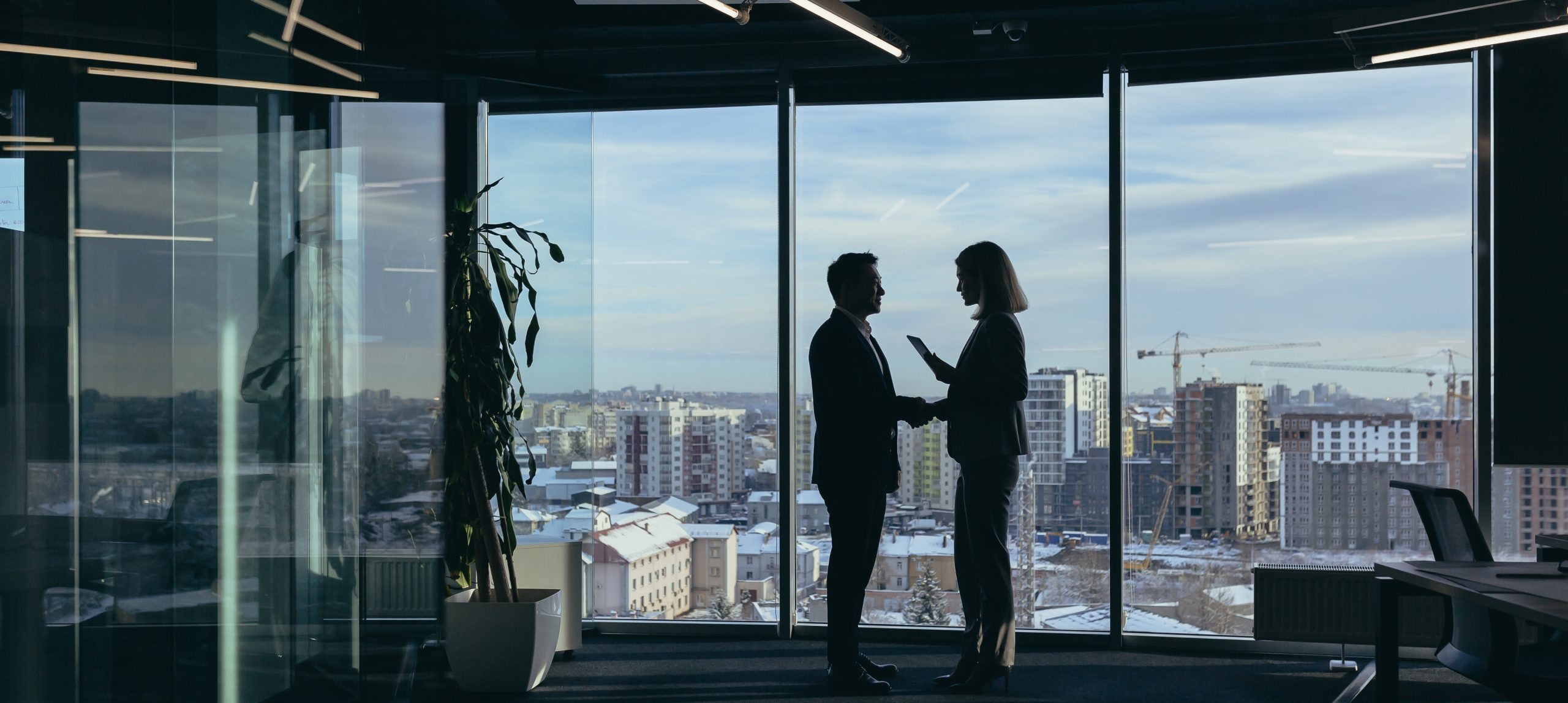 Two professionals in suits shake hands in a glass-walled office with a city skyline visible outside behind them.