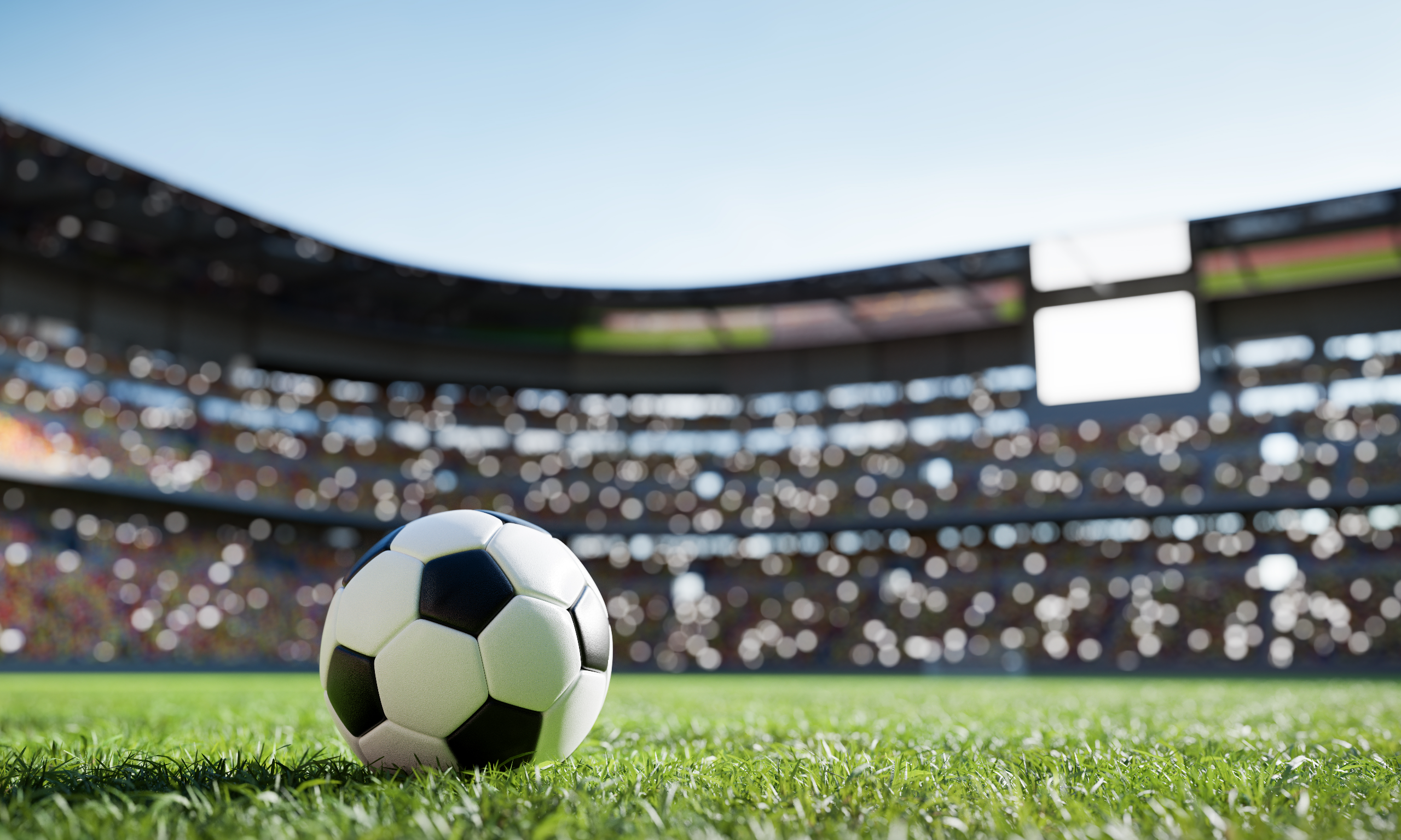 Soccer ball on green grass in a sunlit stadium with a blurred crowd in the background.