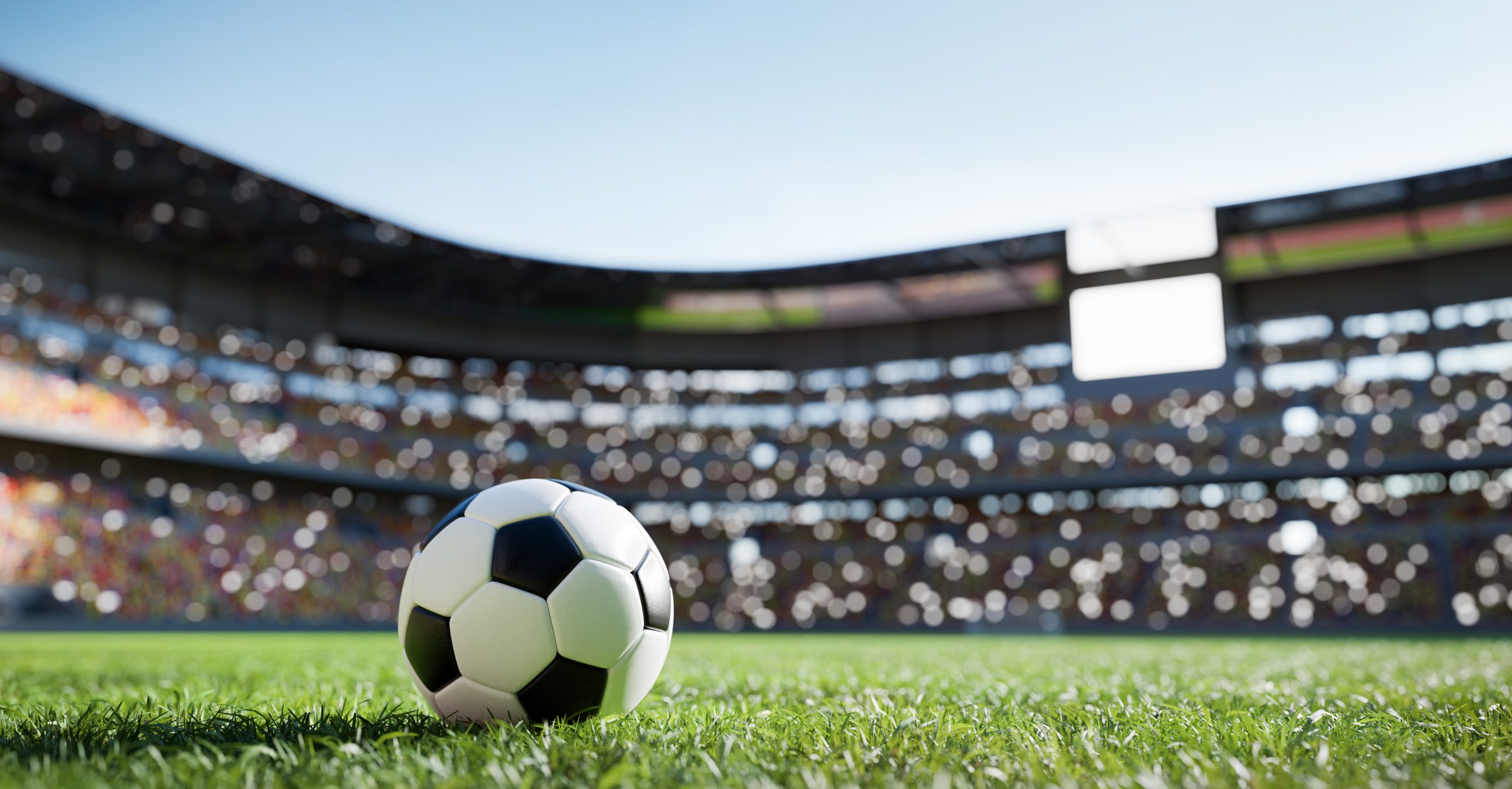 Soccer ball on grass with a blurred stadium crowd in the background, bright blue sky above.